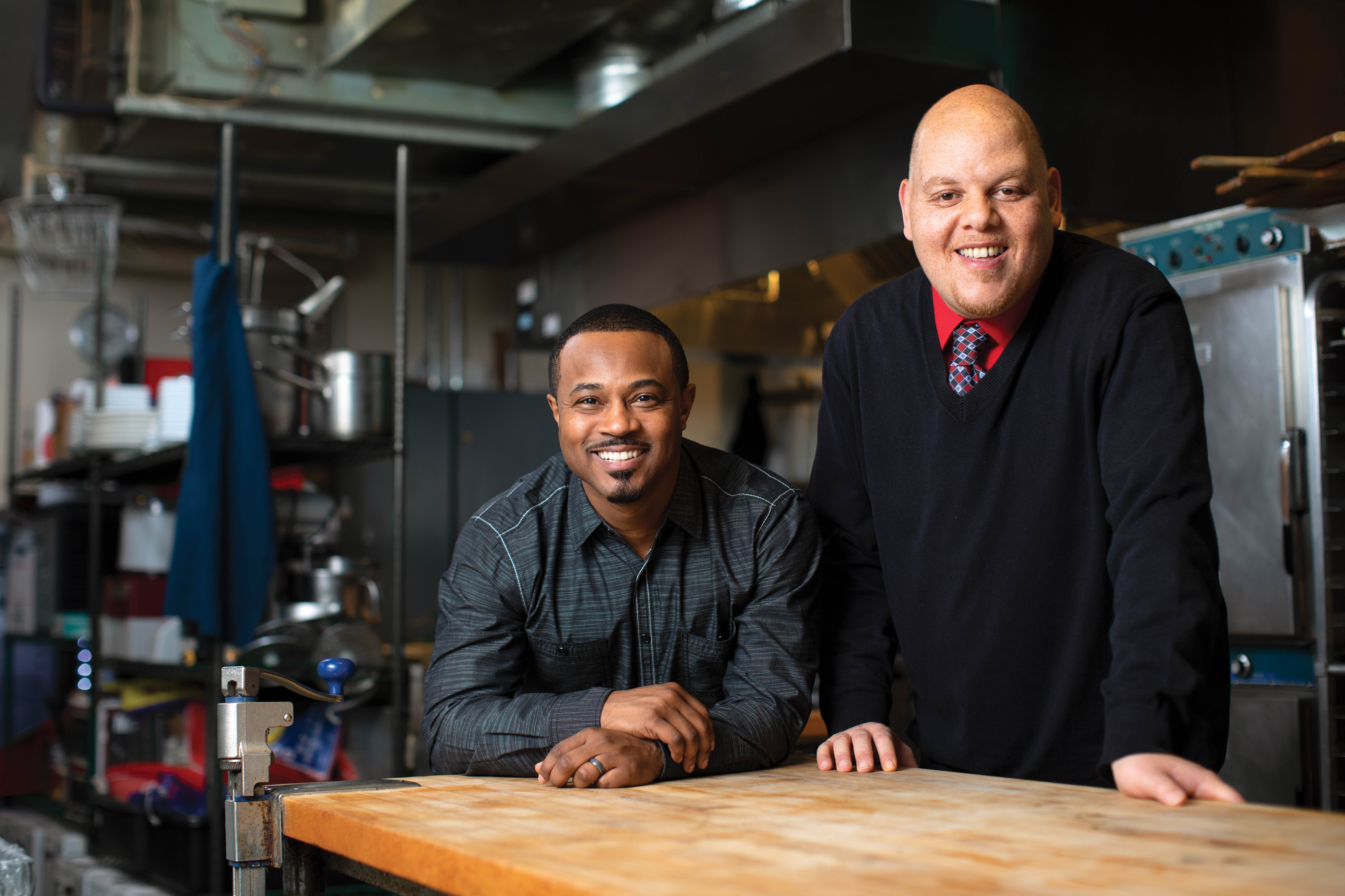 Two men smiling at a work table in an industrial kitchen